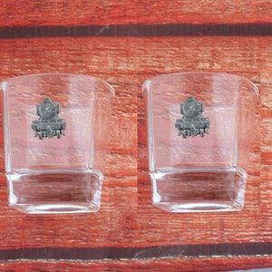 May include: Two clear glass tumblers with a pewter locomotive design on the front. The tumblers are on a wooden surface.