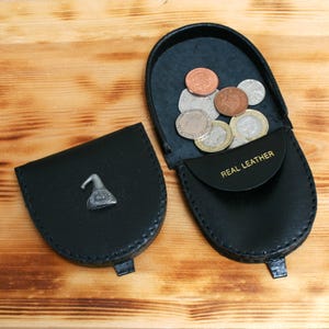 May include: Black leather coin purse with a silver emblem and a snap closure. The open purse reveals several coins. The words "REAL LEATHER" are printed in gold on the inside flap. The purse is on a wooden surface.