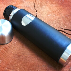 K&ouml;nnte beinhalten: Eine schwarze, lederbezogene Thermoskanne mit silbernem Deckel und einem silbernen Akzent an der Seite. Die Thermoskanne liegt auf einer Holzoberfl&auml;che.