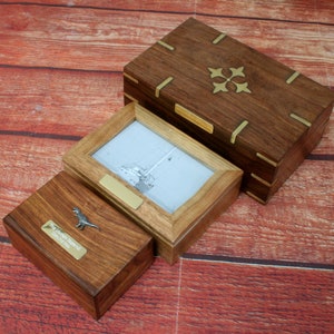 May include: Three wooden boxes with brass accents. The largest box has a decorative brass design on the lid. The middle box has a photo frame with a black and white image of a sailboat. The smallest box has a small metal dinosaur figurine on the lid.