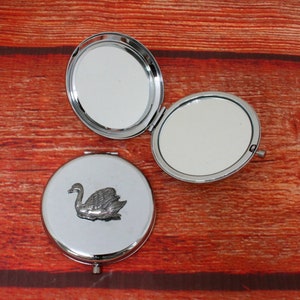 May include: A silver compact mirror with a swan design on the front. The mirror is closed and the swan is facing the right.