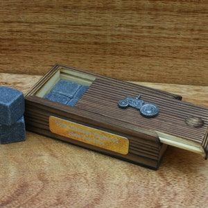May include: A wooden box with a sliding lid, containing grey whiskey stones. The box has a metal tractor charm and a gold plaque with the text "From Lawrence & Ingrid Here's to you Babe".