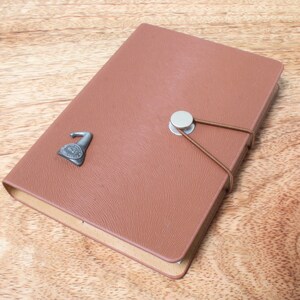 May include: A brown leather-bound journal with a silver clasp and a decorative metal charm. The journal has a textured surface and a simple design.
