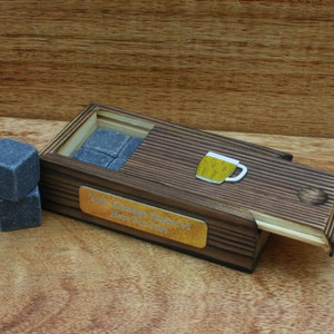 May include: A wooden box with a sliding lid, containing grey whisky stones. The box has a brass plaque with the inscription "Your Mileage May Vary Here's to You Friend". A small beer mug icon is on the lid.