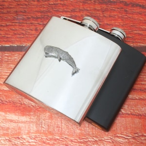 Puede incluir: Dos petacas de acero inoxidable, una plateada y otra negra. La petaca plateada presenta un emblema de ballena. Ambas petacas son rectangulares con tapones de rosca. El fondo es una superficie de madera.