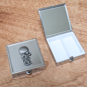 May include: A silver pill box with a hinged lid and a mirror inside. The box has a raised design of a person holding a globe on their head.