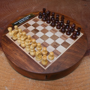 Puede incluir: Un tablero de ajedrez redondo de madera con un diseño de madera marrón y clara. El tablero está abierto y muestra una partida en curso con piezas de ajedrez marrones y blancas. El tablero tiene una placa de latón con el texto "Your Message Engraved Here for Free."