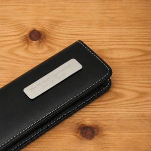 K&ouml;nnte beinhalten: Ein schwarzes Kunstleder-Stiftetui mit wei&szlig;en N&auml;hten und einem silbernen Namensschild. Auf dem Namensschild steht "Your Message Engraved Here Free Of Charge". Das Etui ist geschlossen und liegt auf einer Holzoberfl&auml;che.