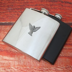 Peut inclure: Une gourde en acier inoxydable argentée et noire avec un motif d'ange. La gourde est posée sur une surface en bois.