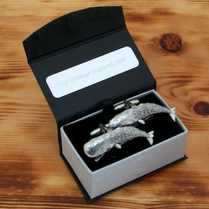以下が含まれることがあります： 黒い箱に白い内側がついた銀製のカフスボタンのセット。カフスボタンはクジラの形をしています。箱には「メッセージを刻印してください」と書かれた白いラベルが付いています。