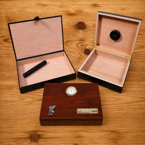 May include: Two open wooden cigar humidors with light wood interiors and black exteriors, one containing a black cylindrical humidifier. A third closed dark wood humidor features a round analog hygrometer, a silver fairy emblem, and a gold plaque.
