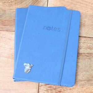 May include: Two blue notebooks with a koala sticker on the front. The notebooks have a black elastic band and the word "notes" printed on the front.