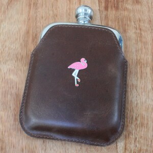 Könnte beinhalten: Eine braune Lederflasche mit silbernem Verschluss und einem pinkfarbenen Flamingo-Motiv.