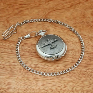 Könnte beinhalten: Silberne Taschenuhr mit Kette und einem detaillierten Bild eines Vintage-Flugzeugs auf dem Zifferblatt.