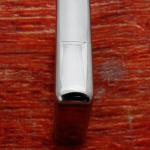 Puede incluir: Un encendedor plateado con un acabado liso y pulido. El encendedor tiene una forma rectangular con bordes redondeados y una peque&ntilde;a abertura negra en la parte inferior.