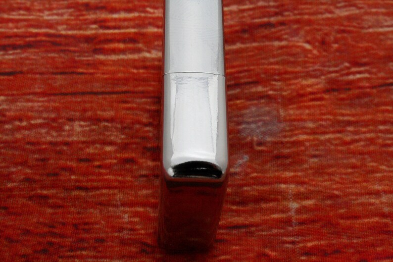 Puede incluir: Un encendedor plateado con un acabado liso y pulido. El encendedor tiene una forma rectangular con bordes redondeados y una peque&ntilde;a abertura negra en la parte inferior.