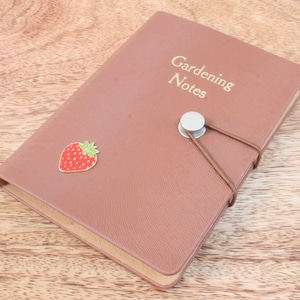 Peut inclure: Un carnet relié en cuir marron avec une inscription dorée qui dit "Gardening Notes". Une petite épingle à fruits rouges en forme de fraise est fixée à la couverture.