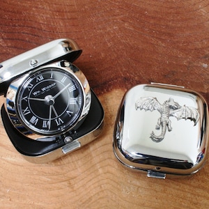 Puede incluir: Un reloj de bolsillo plateado con una esfera negra y números romanos. El reloj está en una caja plateada con un diseño de dragón en la tapa. El reloj está sobre una superficie de madera.