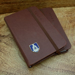 May include: Two brown leather-bound notebooks with elastic closures. The top notebook has a gold-colored emblem with a blue center and the word "NOTES" embossed on the cover.