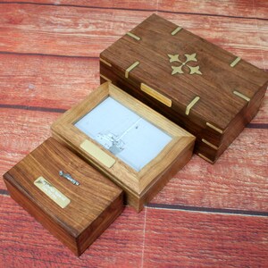 May include: Three wooden boxes with brass accents. The largest box has a decorative brass design on the lid. The middle box has a picture frame with a black and white photo of a sailboat. The smallest box has a brass plaque with the text "First Mate" on it.