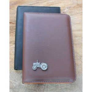 May include: A brown leather notebook with a small metal tractor charm. The notebook is closed and has a black leather cover behind it.