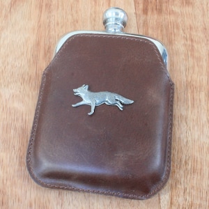 Puede incluir: Una petaca de cuero marrón con un diseño de zorro plateado en la parte delantera.