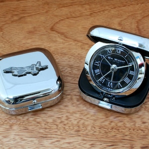 May include: A silver-coloured travel alarm clock with a black face and Roman numeral hour markers. The clock is in a silver-coloured case with a hinged lid. The case has a small, silver-coloured airplane design on the top.