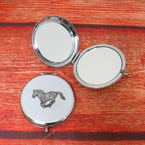 May include: A silver compact mirror with a running horse design on the front. The mirror is open, revealing a silver interior with two mirrors.