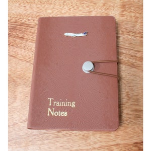 May include: A brown leather notebook with the words "Training Notes" in gold lettering. A silver airplane charm is on the cover. The notebook is closed with a circular clasp and elastic cord.