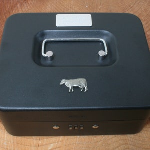 Puede incluir: Caja de metal negra con una cerradura de combinación y una figura de vaca plateada en la tapa. La tapa tiene una manija plateada.
