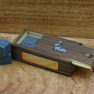 Puede incluir: Una caja de madera con tapa deslizante, grabada con el texto "Your Message Here For Free". La caja contiene piedras de whisky grises y una figurita de conejo de plata.