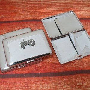 May include: Two silver cigarette cases with a tractor design on the front. The cases are open and show the interior.