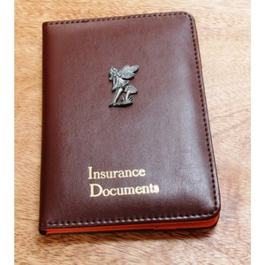 Può includere: Un portadocumenti per assicurazioni in pelle marrone con una piccola statuetta di fata argentata sul davanti. Il portadocumenti ha le parole "Insurance Documents" in rilievo in lettere dorate.