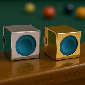 May include: Two metallic cube-shaped billiard chalk holders, one silver and one gold, each with a teal chalk insert. The holders are on a wooden surface, with billiard balls in the background. The holders have a small latch on the side.