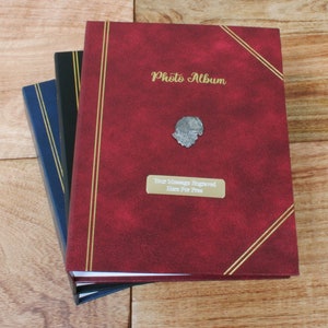 May include: Three photo albums with gold trim. The top album is red with the words "Photo Album" in gold lettering. The middle album is black and the bottom album is blue.