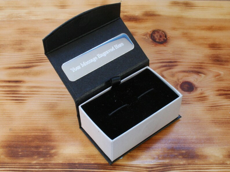 May include: A black cufflink box with a white interior and a silver plate on the top that says "Your message engraved here".