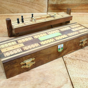 May include: A wooden cribbage board with a hinged lid and a small wooden box containing a cribbage board. The board has a green label with the text "Cribbage" on it. The box has a brass latch and a small metal emblem on the side.