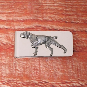 May include: A polished silver money clip with a sculpted dog design. The dog is depicted in a standing position, with intricate detailing. The clip's surface is highly reflective, catching the light.