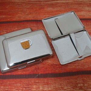 May include: Silver cigarette case with a hinged lid and a small beer mug design on the front.
