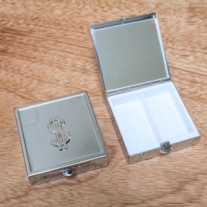 Może przedstawiać: Srebrne pudełko na tabletki z nadrukiem znaku dolara z przodu i lustrem w środku. Pudełko na tabletki ma dwa przedziały.