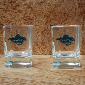 May include: Two clear glass shot glasses with a pewter emblem on the front. The emblem is a map of the Isle of Wight with the text "Isle of Wight" underneath.