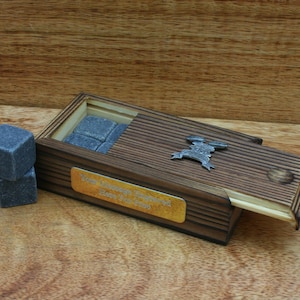 Puede incluir: Una caja de madera con tapa abatible, que contiene piedras de whisky grises. La caja tiene una placa dorada con la inscripción "You Always Brighten Our Day, Here's to You!" y un amuleto de metal plateado.