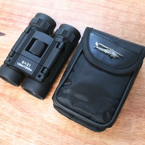 Könnte beinhalten: Schwarzes Fernglas mit Tragetasche. Das Fernglas hat eine Vergrößerung von 8x21 und ein Sichtfeld von 128m/1000m. Die Tragetasche hat ein silbernes Helikopter-Emblem.