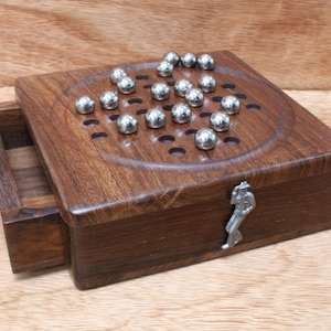 Puede incluir: Un tablero de juego de madera con un cajón y una figurita de metal. El tablero tiene una depresión circular con agujeros y bolas de metal plateadas.
