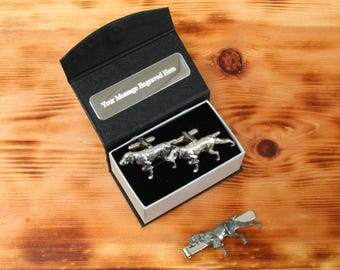 Puede incluir: Un juego de gemelos de plata y un clip para corbata con un diseño de perro de caza. Los gemelos están en una caja negra con una placa de plata que dice "Su mensaje grabado aquí".