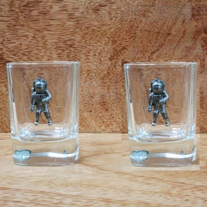 May include: Two clear glass shot glasses with a pewter astronaut figurine on the front of each glass.