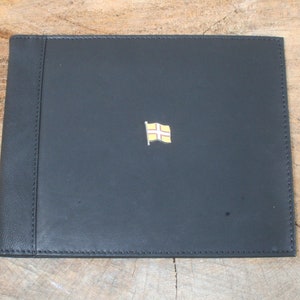 Könnte beinhalten: Schwarzes Lederfotoalbum mit einer kleinen goldenen Flagge der Kanalinseln auf dem vorderen Umschlag.