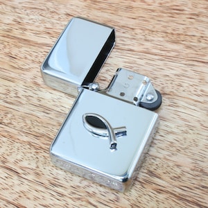 May include: A silver lighter with a black fish symbol on the front. The lighter is open and the lid is laying next to it.