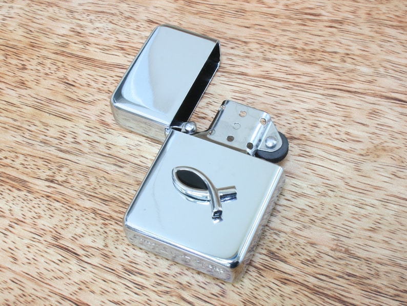 May include: A silver lighter with a black fish symbol on the front. The lighter is open and the lid is laying next to it.