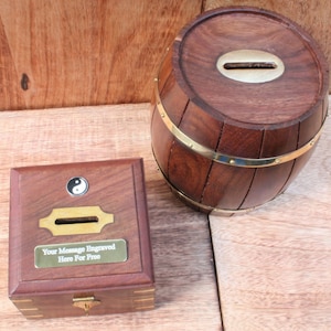 Puede incluir: Dos cajas de madera para guardar dinero, una con forma de barril y la otra con forma de caja cuadrada. El barril tiene bandas de latón alrededor y una ranura en la parte superior. La caja cuadrada tiene un cierre de latón y una ranura en la parte delantera. Ambas cajas tienen una placa de latón con el texto "Su mensaje grabado aquí gratis".